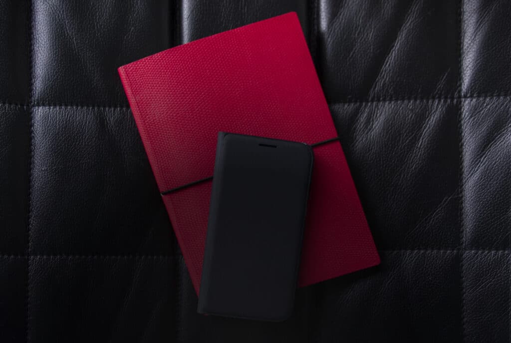 A mobile phone sits on a pink notebook which sits on a leather banquette
